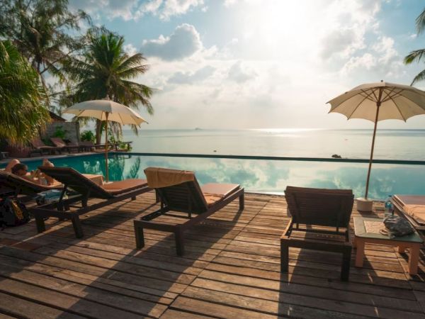 Poolside view with lounge chairs, umbrellas, and palm trees under a sunny sky reflected in the water, creating a serene atmosphere.
