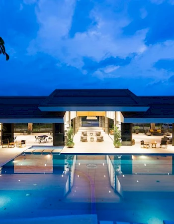 A luxurious building with an illuminated pool reflecting the evening sky, surrounded by seating areas under a clear blue ambiance.