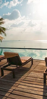A serene poolside scene with lounge chairs, umbrellas, and a scenic view of palm trees and water under a partly cloudy sky.