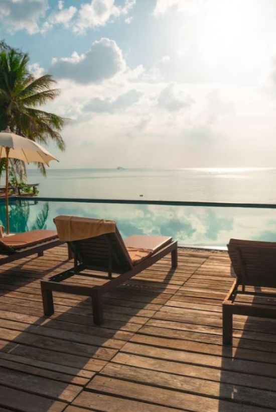 A serene poolside setting with lounge chairs, umbrellas, and a view of the ocean under a cloudy sky and palm trees.