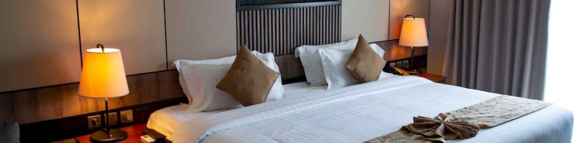 A neatly arranged hotel room with a large bed, decorative pillows, side lamps, and curtains, creating a cozy and inviting atmosphere.