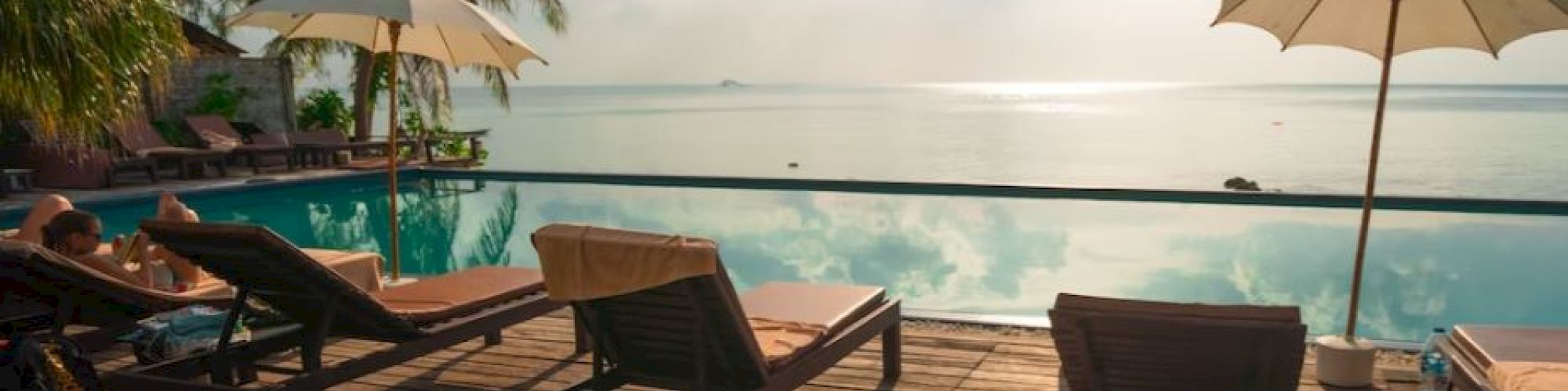 Poolside scene with lounge chairs and umbrellas on a wooden deck facing a calm sea and partly cloudy sky, creating a tranquil setting.