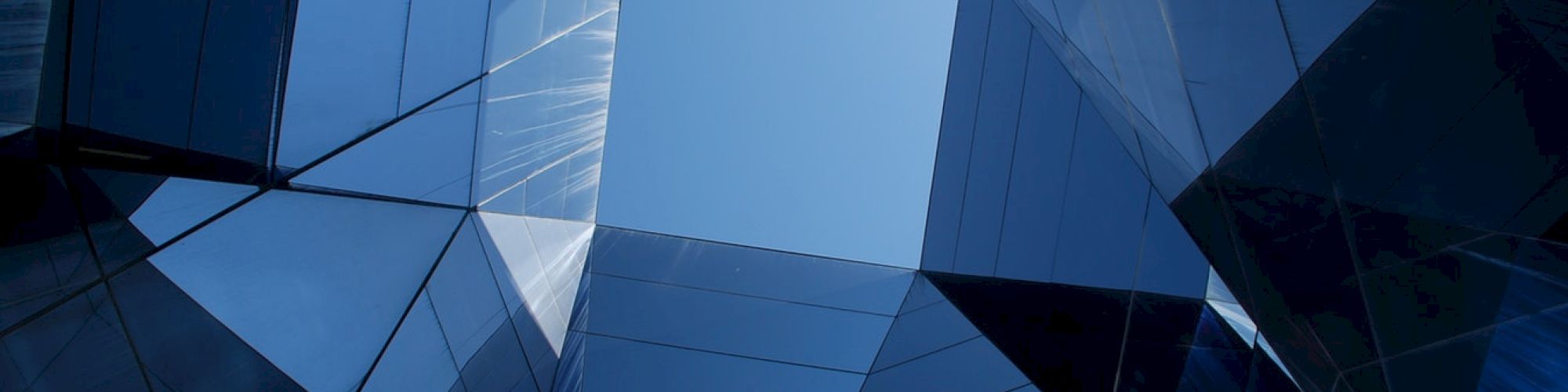 A view looking up through geometric, reflective glass structures with a clear blue sky visible in the center.