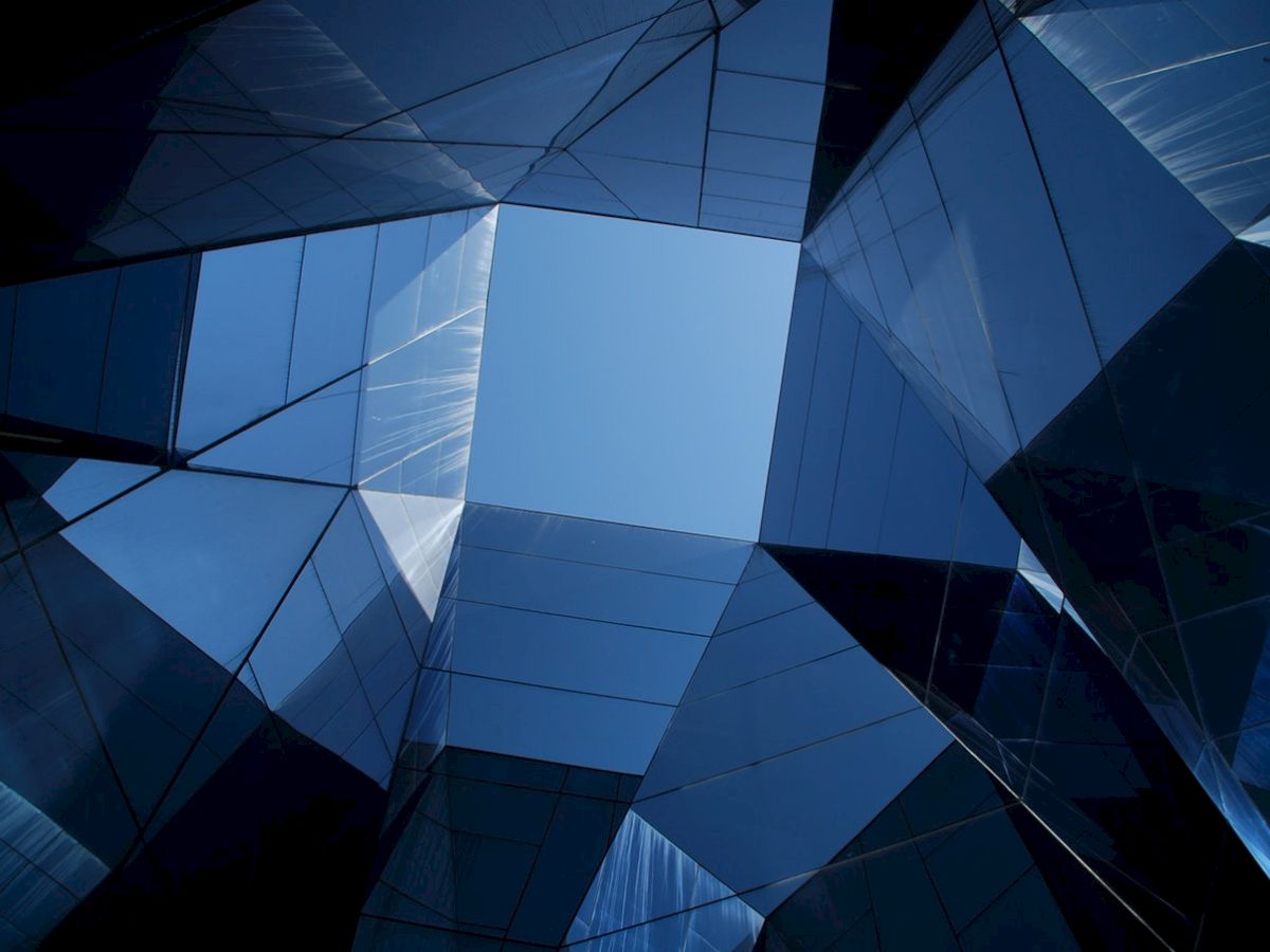 The image shows a modern architectural structure with angular glass panels, reflecting a blue sky in a geometric pattern.