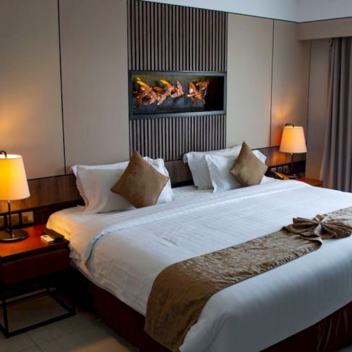 A cozy hotel room features a large bed with brown accents, decorative pillows, bedside lamps, and curtains by a window.
