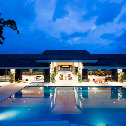 A modern building at night with a large illuminated swimming pool and seating areas, framed by a deep blue sky.