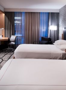 The image shows a modern hotel room with two beds, a large TV, a desk, and stylish decor featuring patterned wallpaper.