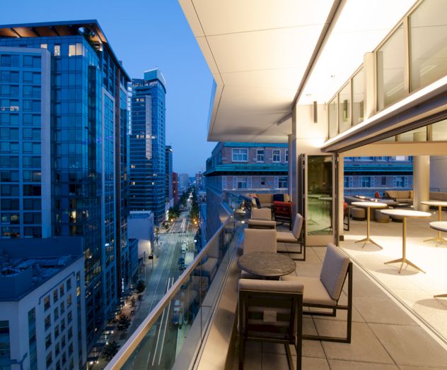 A modern cityscape with tall buildings and a stylish terrace featuring outdoor seating, overlooking the street below at dusk.