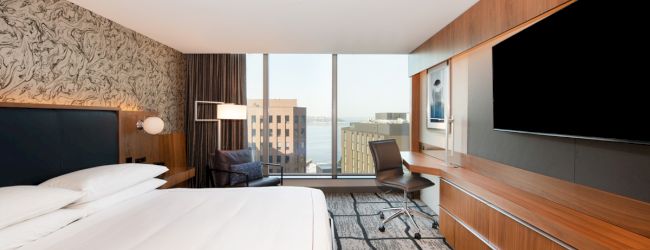 A modern hotel room with a large bed, desk, chair, TV, and a window view of the cityscape and water.