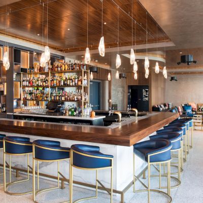 A modern bar with a polished counter, blue chairs, hanging lights, and a well-stocked liquor shelf, surrounded by comfortable seating.