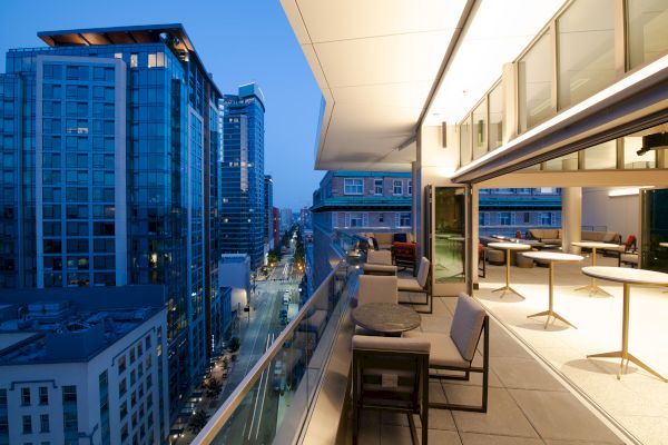 A modern balcony overlooks a city street, with surrounding skyscrapers lit up at twilight. The space has tables and seating arrangements.
