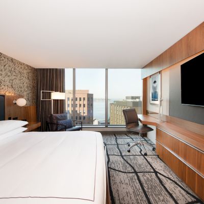 A modern hotel room featuring a large bed, wall-mounted TV, desk, chair, and city view through a large window.
