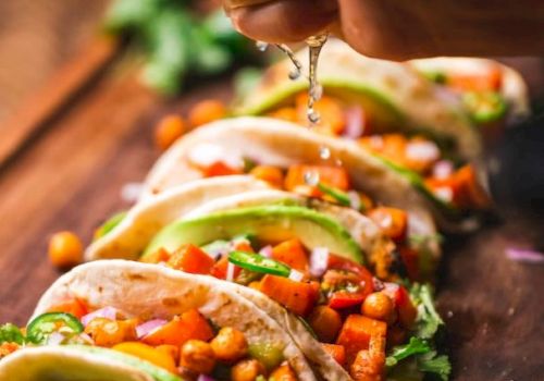 A hand squeezes lime over tacos filled with vegetables, chickpeas, and garnished with avocado slices on a wooden board.
