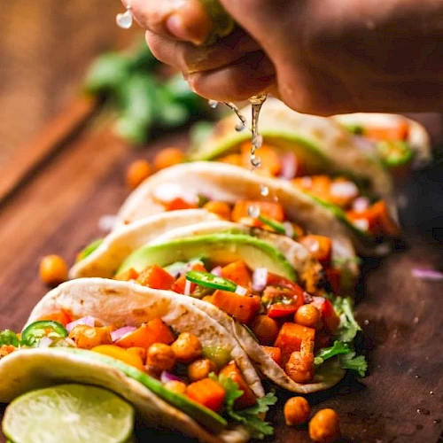 A hand squeezes lime over tacos filled with vegetables, chickpeas, and garnished with avocado slices on a wooden board.