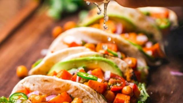 A hand squeezes lime over tacos filled with vegetables, chickpeas, and garnished with avocado slices on a wooden board.
