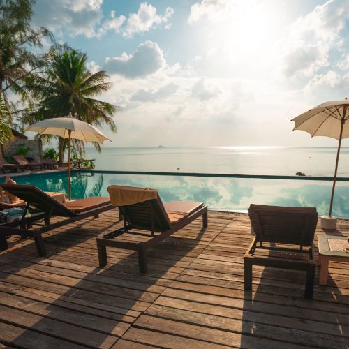Three lounge chairs under umbrellas overlook an infinity pool leading to a serene ocean view, with palm trees and a cloudy sky above.