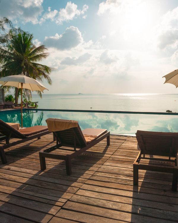 Three lounge chairs under umbrellas overlook an infinity pool leading to a serene ocean view, with palm trees and a cloudy sky above.