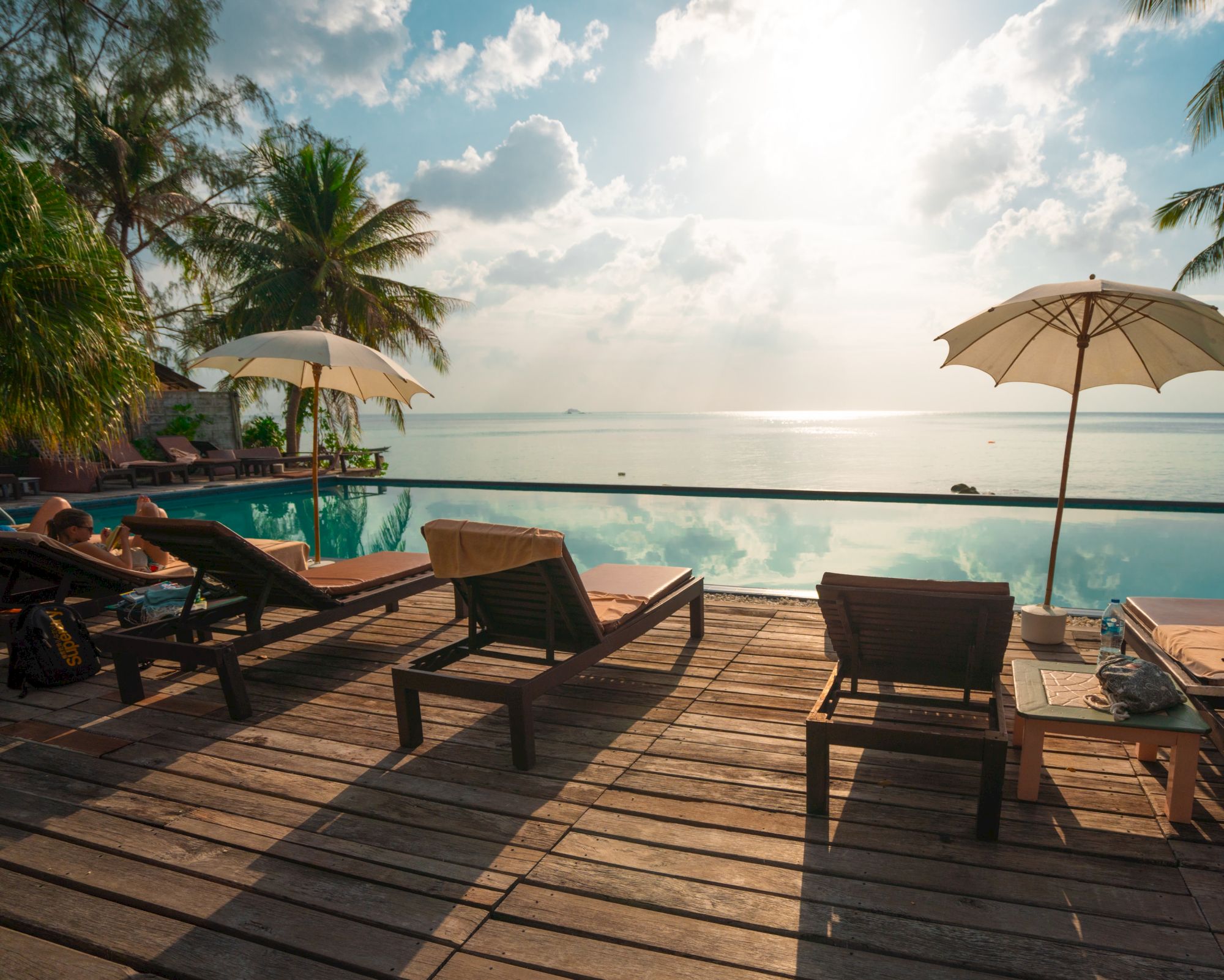 Lounge chairs and umbrellas by an infinity pool overlook the ocean, surrounded by palm trees under a partly cloudy sky.