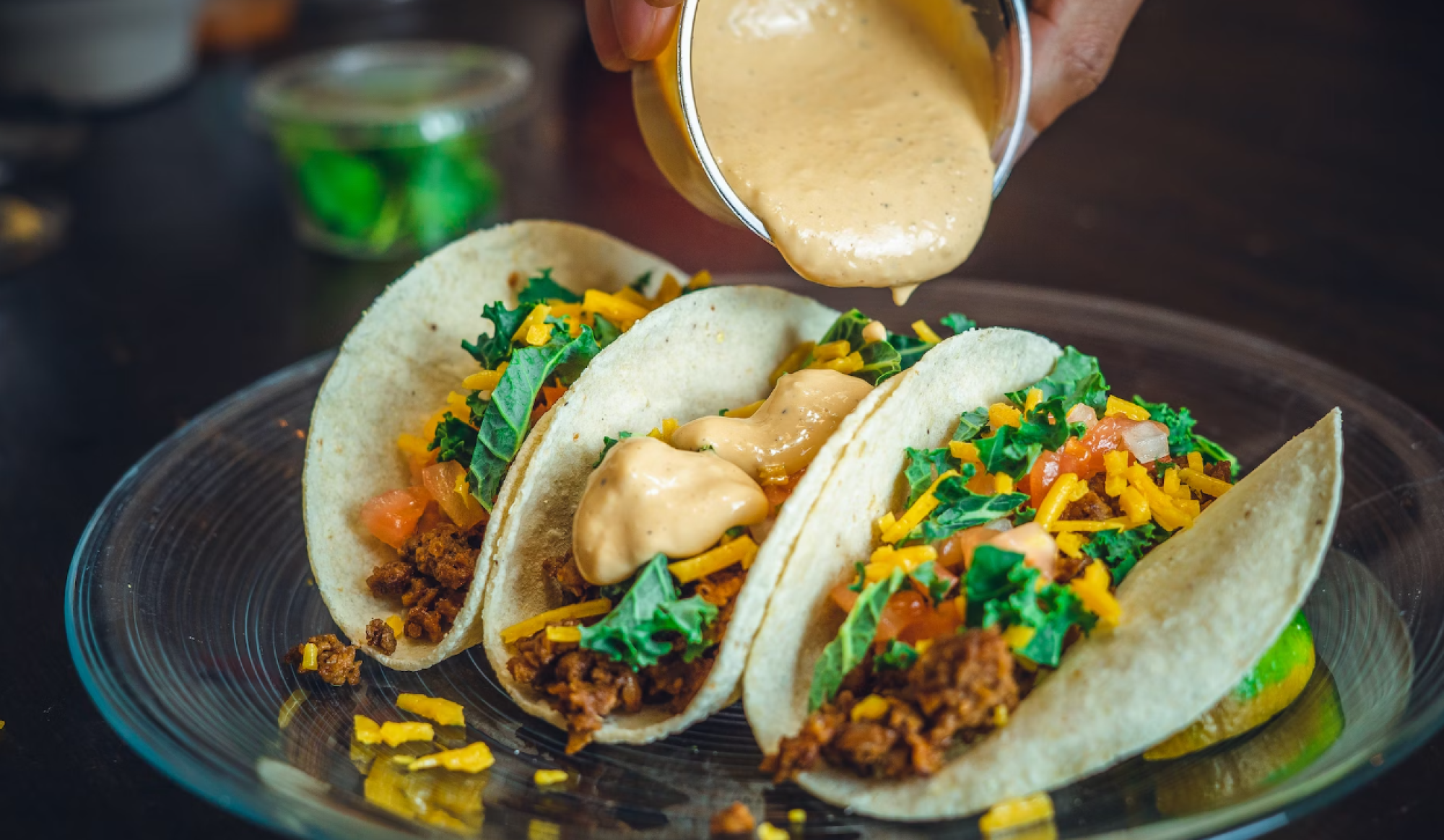 Three tacos with meat, cheese, lettuce, and tomatoes on a plate. Sauce is being poured over them, enhancing their flavor.