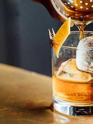 A hand pours a brown cocktail into a glass with ice and an orange peel garnish on a bar top.