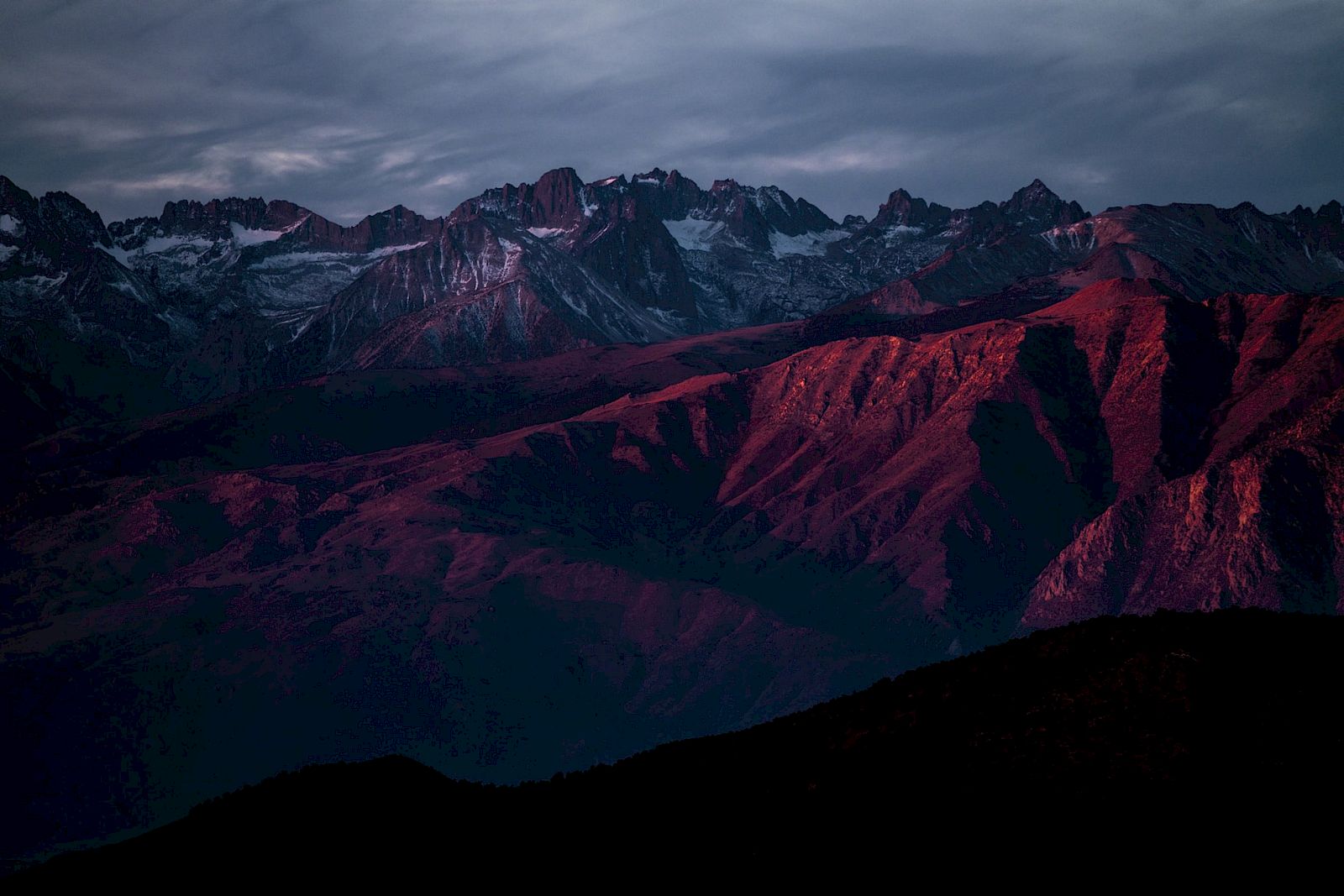 A mountainous landscape with rugged peaks and slopes is bathed in dramatic, reddish light during sunset or sunrise, under a cloudy sky.