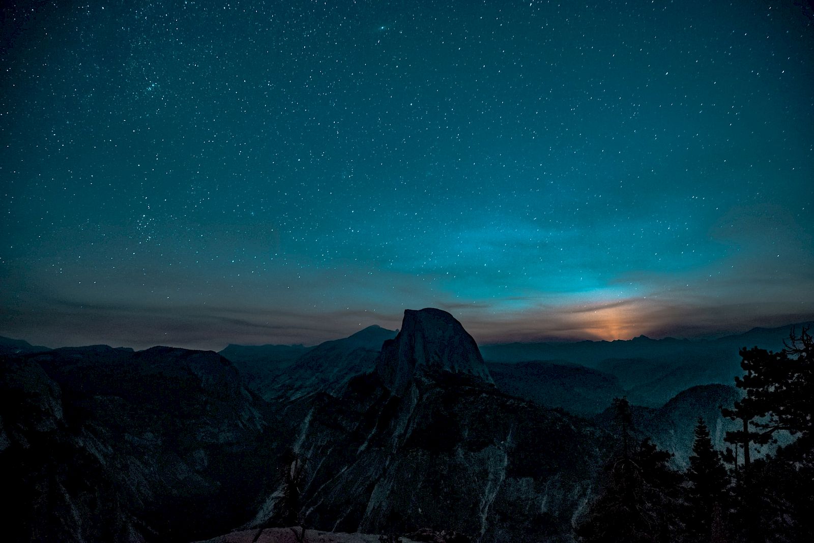 A starry night sky with a silhouetted mountain range and a tree line in the foreground, illuminated by a distant light source.