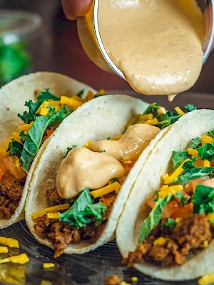 Three tacos filled with seasoned ground meat, shredded cheese, greens, and tomatoes, being topped with a creamy sauce on a plate.