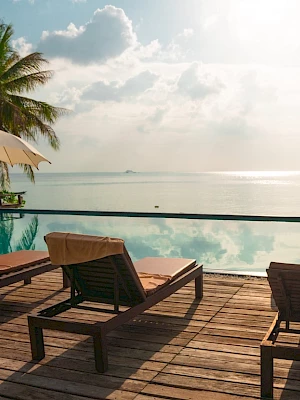 Lounge chairs and umbrellas by a poolside with an ocean view, surrounded by palm trees under a partly cloudy sky.