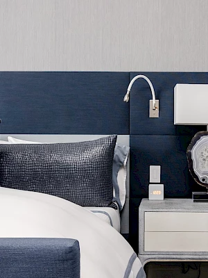 A modern bedroom with a blue padded headboard, white bed, two pillows, a bedside table, lamp, and reading light on a grey wall.