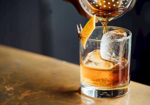 A drink is being poured into a glass with ice and a citrus peel garnish, resting on a wooden surface.