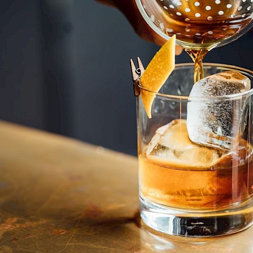 A drink is being poured into a glass with ice and a citrus peel garnish, resting on a wooden surface.