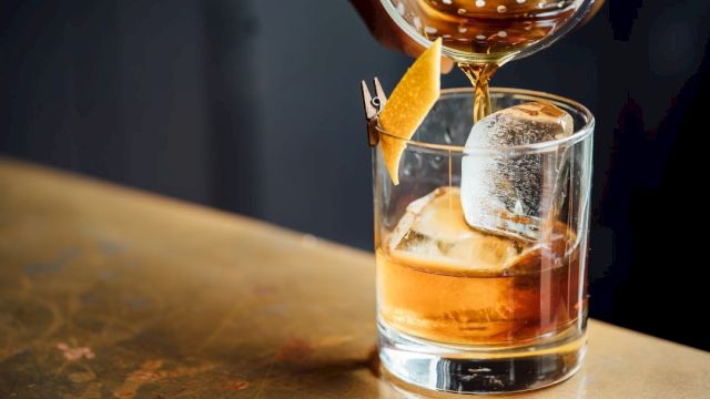 A drink is being poured into a glass with ice and a citrus peel garnish, resting on a wooden surface.