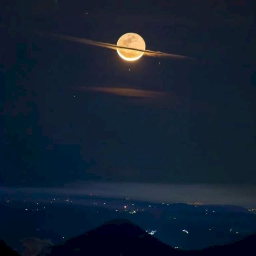 A full moon with clouds creating a "Saturn-like" appearance in the night sky, overlooking a landscape with glowing city lights below.