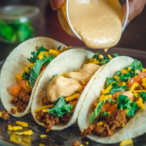 Three tacos with seasoned meat, cheese, kale, and tomatoes, topped with sauce being poured, served on a plate.