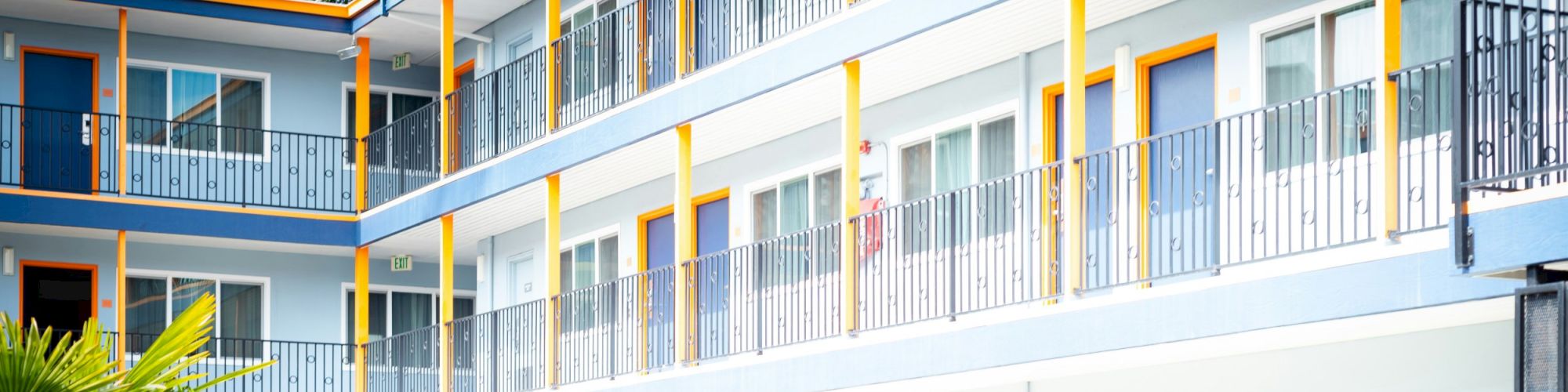 The image shows a three-story building with balconies, blue and yellow details, and glass doors. There's a palm tree in the foreground.