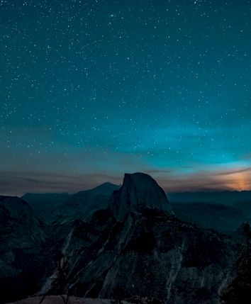 A nighttime landscape with a starry sky, a mountain silhouette, and a soft glow on the horizon resembling moonrise or sunrise.