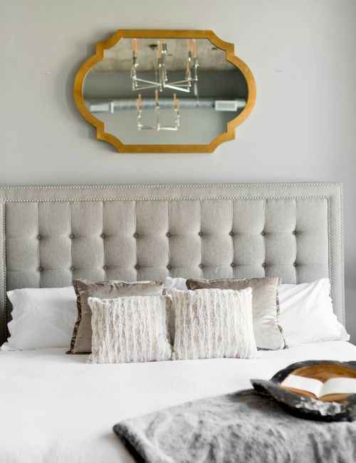 A stylish bedroom features a tufted headboard, decorative pillows, lamps on side tables, a unique mirror, and an open book on the bed.