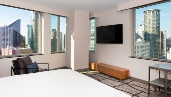 A modern hotel room features large windows with city views, a wall-mounted TV, a bed, a small table, and a lounge chair with a pillow.
