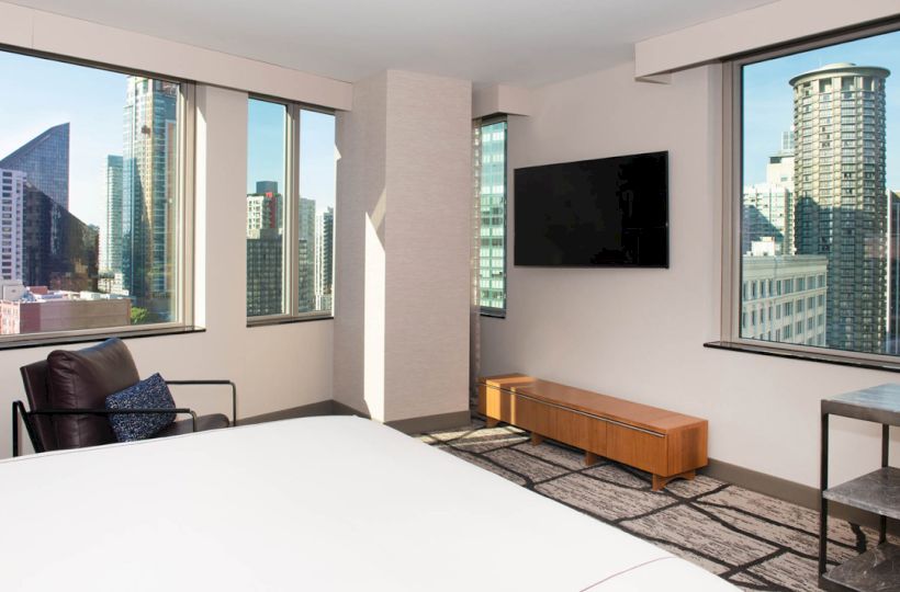 A modern hotel room features large windows with city views, a wall-mounted TV, a bed, a small table, and a lounge chair with a pillow.
