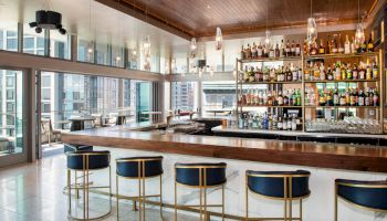 The image shows a modern bar with elegant stools, a wide selection of bottles, and cityscape views from large windows.