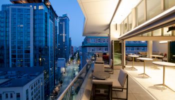The image shows a modern balcony overlooking a cityscape with tall buildings. Tables and chairs are on the balcony, city lights are visible.