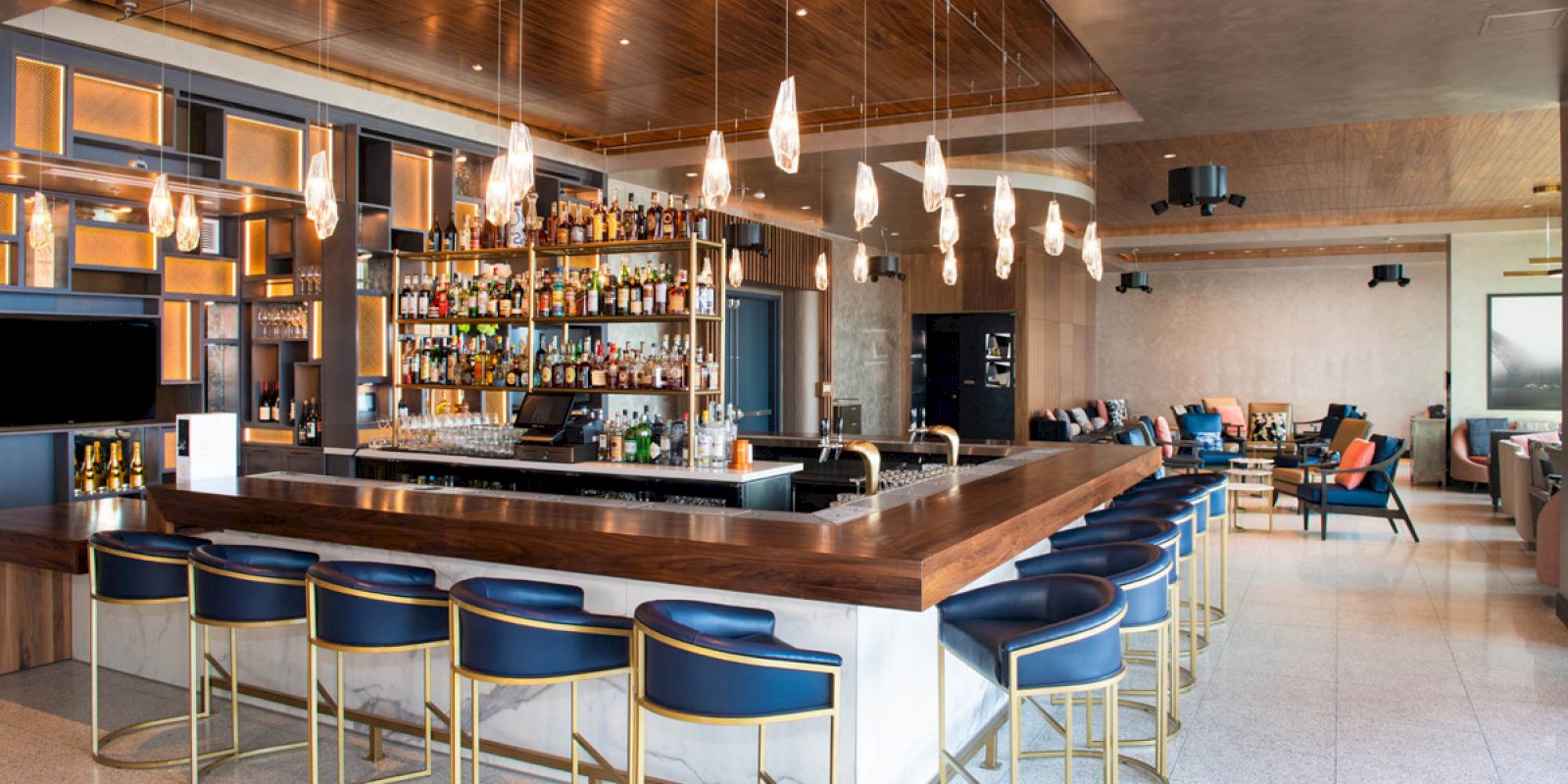 The image shows a modern bar with a central counter, bar stools, overhead lights, and a variety of bottles on display in the background.