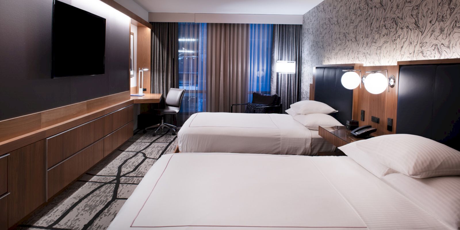 The image shows a modern hotel room with two double beds, a flat-screen TV, a desk with a chair, and stylish lighting and decor.