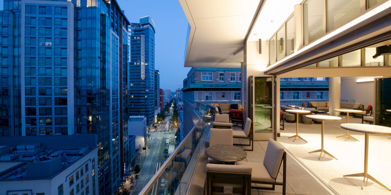A cityscape viewed from a modern balcony at dusk, featuring outdoor tables and chairs, and tall buildings lining a lit street.