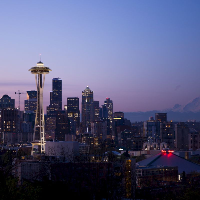 Seattle skyline at dusk featuring the Space Needle with distant Mount Rainier in the background under a clear purple-blue sky.