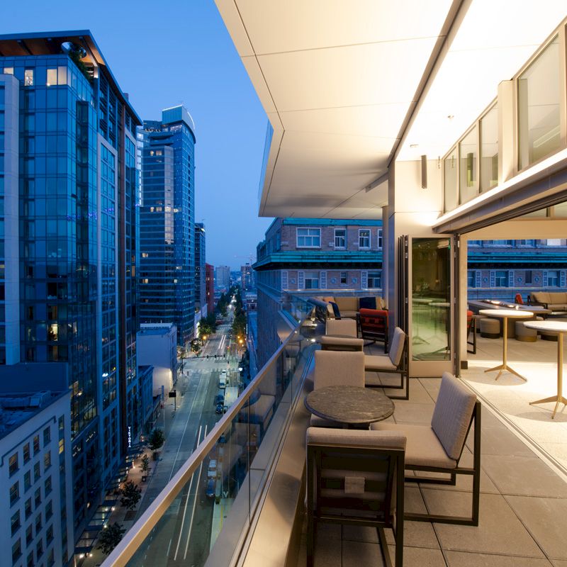 A modern rooftop terrace with seating overlooks a cityscape of tall buildings at dusk, featuring a mix of glass and illuminated interiors.