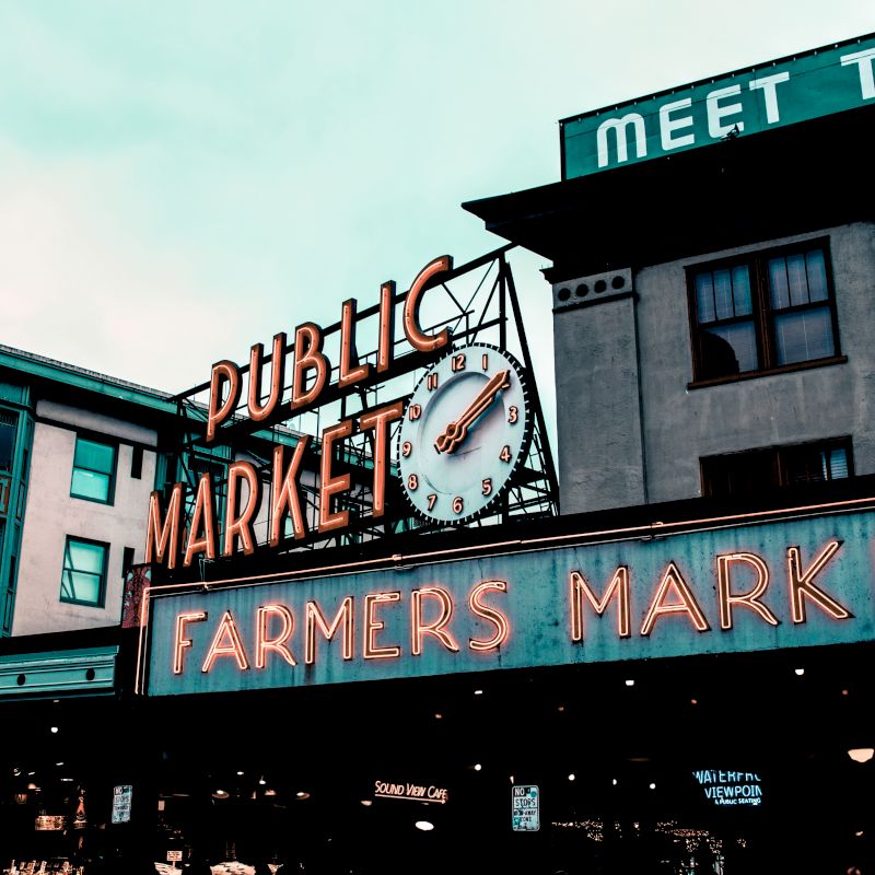 The image shows the iconic sign for a public farmers market, with neon lights and a clock, set against urban buildings in the background.