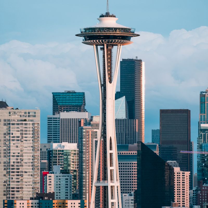 The image shows the Space Needle in Seattle, surrounded by skyscrapers and buildings, with a partly cloudy sky in the background.