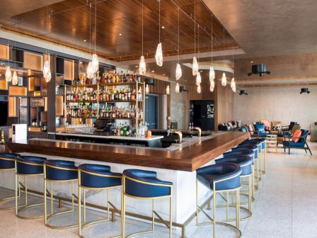 A modern bar with a countertop, barstools, hanging lights, and a stocked liquor shelf in a spacious room with additional seating.