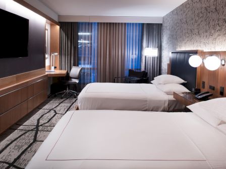 The image shows a modern hotel room with two beds, a desk, and a TV. The room features contemporary decor and a large window.
